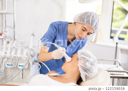 Female cosmetologist performing high frequency ultrasonic facial procedure on woman Female cosmetologist performing high frequency ultrasonic facial procedure on woman 122159016