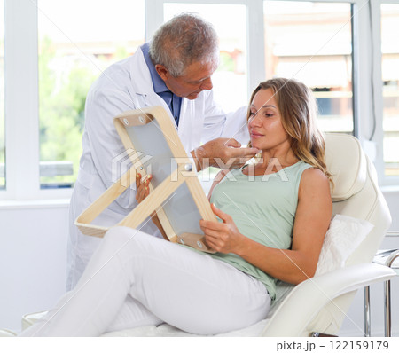 Woman looking at mirror while elderly cosmetologist showing results of facial procedure Woman looking at mirror while elderly cosmetologist showing results of facial procedure 122159179