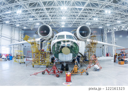 Front view of the white transport airplane in the hangar. Aircraft under maintenance. Checking mechanical systems for flight operations Front view of the white transport airplane in the hangar. Aircraft under maintenance. Checking mechanical systems for flight operations 122161323