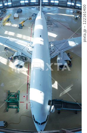 Top view of a white passenger jet plane in the hangar. Airplane under maintenance. Checking mechanical systems for flight operations Top view of a white passenger jet plane in the hangar. Airplane under maintenance. Checking mechanical systems for flight operations 122161400