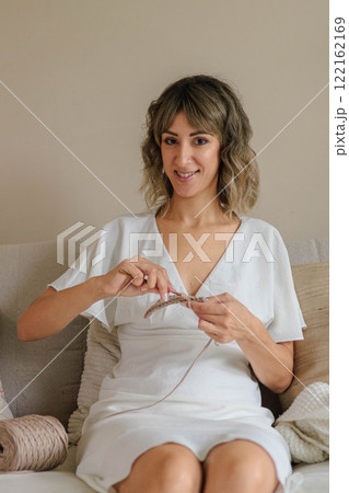 Young beautiful woman sits on the sofa and crochets from beige yarn. The girl enjoys the hobby. Young beautiful woman sits on the sofa and crochets from beige yarn. The girl enjoys the hobby. 122162169