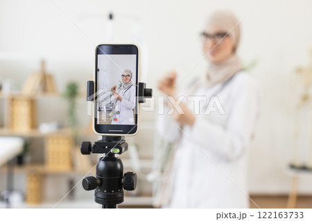 Middle-eastern adult female doctor recording video lesson on medical procedure. Demonstrates how to use a dropper correctly via smartphone. Educational content for healthcare professionals. 122163733