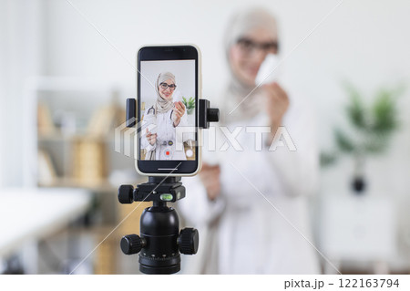 Muslim woman in hijab demonstrating medicine bottles while recording video. Young adult female creating educational content on healthcare. Professional setting suggests engaging online audience. Muslim woman in hijab demonstrating medicine bottles while recording video. Young adult female creating educational content on healthcare. Professional setting suggests engaging online audience. 122163794