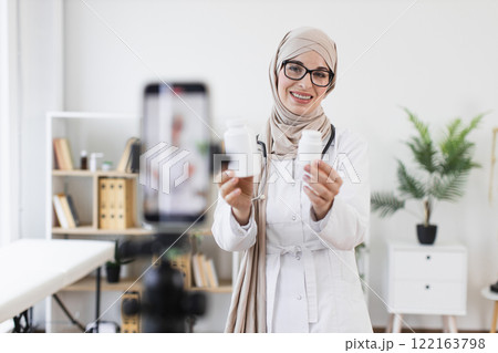 Muslim woman doctor with glasses recording video about medical treatment using phone. Middle-aged female sharing knowledge about medicine in modern office setting. 122163798