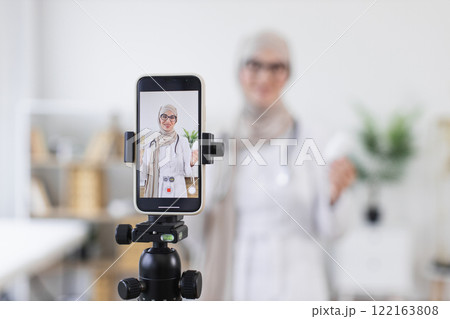 Muslim woman recording video lesson on medicine with smartphone in modern setting. Middle Eastern adult shares treatment tips online, combining traditional and digital methods for health awareness. Muslim woman recording video lesson on medicine with smartphone in modern setting. Middle Eastern adult shares treatment tips online, combining traditional and digital methods for health awareness. 122163808