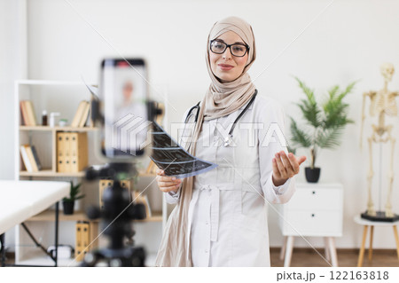 Muslim woman doctor recording educational video with x-ray, highlighting medical treatment. Female professional showcases modern healthcare approach, creating online content for patient education. 122163818