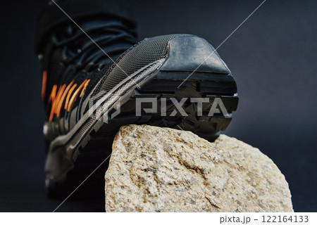 Close-up of waterproof black hiking shoe with orange details on small rock. Rugged trekking boot on dark background. Footwear for exploration and outdoor activities 122164133