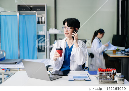 Confident young male doctor in white medical uniform sit at desk working on computer 122165020