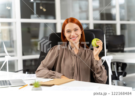Pretty smiling ginger young woman with apple in hands sitting in the office Pretty smiling ginger young woman with apple in hands sitting in the office 122168484
