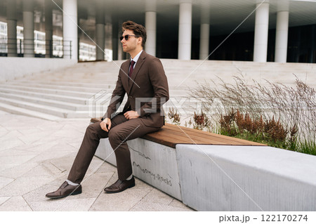 Photogenic businessman in brown suit and sunglasses sitting confidently on city bench, exuding professionalism and success in modern urban setting. Handsome bearded business man posing outdoors. Photogenic businessman in brown suit and sunglasses sitting confidently on city bench, exuding professionalism and success in modern urban setting. Handsome bearded business man posing outdoors. 122170274