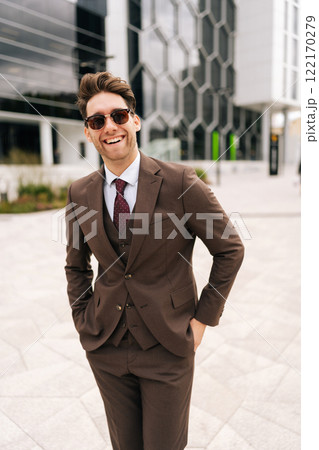 Vertical portrait of cheerful businessman wearing stylish sunglasses and suit standing posing at city, smiling looking at camera, holding hands in pockets, exuding professionalism and leadership. Vertical portrait of cheerful businessman wearing stylish sunglasses and suit standing posing at city, smiling looking at camera, holding hands in pockets, exuding professionalism and leadership. 122170279