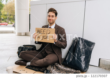 Positive millennial manager in suit holding cardboard sign with inscription need job looking for work after being fired. Homeless man searching job sitting on urban street, smiling looking at camera Positive millennial manager in suit holding cardboard sign with inscription need job looking for work after being fired. Homeless man searching job sitting on urban street, smiling looking at camera 122170307