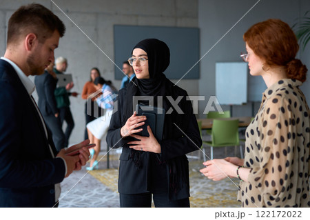 A diverse group of business professionals engages in a conversation during a work break in the office, with their colleagues visible in the background, fostering collaboration and connection 122172022