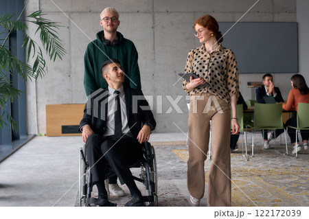 A diverse team of business colleagues, including a director in a wheelchair, engages in a collaborative discussion as they move together through a modern office space 122172039