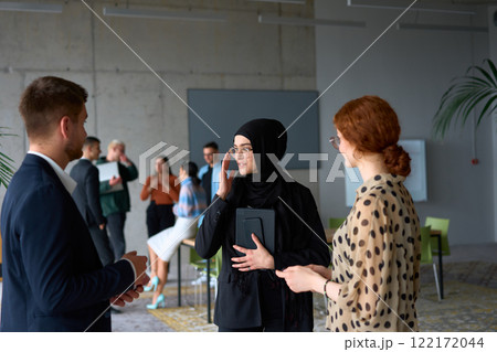 A diverse group of business professionals engages in a conversation during a work break in the office, with their colleagues visible in the background, fostering collaboration and connection 122172044