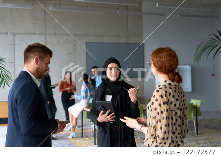 A diverse group of business professionals engages in a conversation during a work break in the office, with their colleagues visible in the background, fostering collaboration and connection 122172327