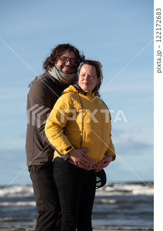A couple stands embraced on a sandy beach on a chilly autumn day, the serene ocean stretching behind them as they share a quiet moment of love and warmth amidst the cool breeze and peaceful 122172683
