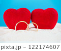 Two red velvet hearts on a beach sand background with a blue sky, accompanied by golden wedding rings. Perfect for themes of Valentine's Day, weddings, or marriage proposals. The image provides space 122174607