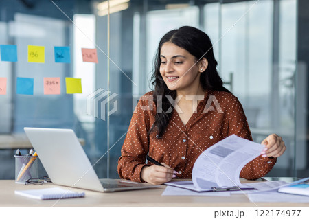 Young successful businesswoman working with documents inside the office. Office worker holding document in hands, filling out tax forms and reports using laptop. Young successful businesswoman working with documents inside the office. Office worker holding document in hands, filling out tax forms and reports using laptop. 122174977