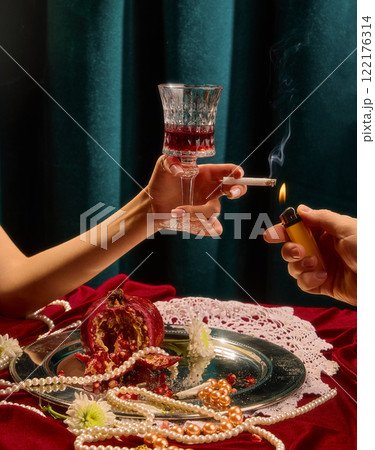 Female hand holding wine glass and cigarette, surrounded by split pomegranate, pearls, and white flowers on silver tray against green velvet background. 122176314