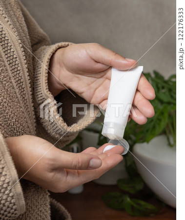 Woman in brown bathrobe applying cream on fingers and holding cosmetic tube in other hand, mockup Woman in brown bathrobe applying cream on fingers and holding cosmetic tube in other hand, mockup 122176333
