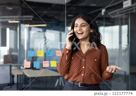 Successful and happy office worker talking on the phone while standing near the window inside the office building. Satisfied businesswoman at workplace communicating with colleagues and clients. 122177774