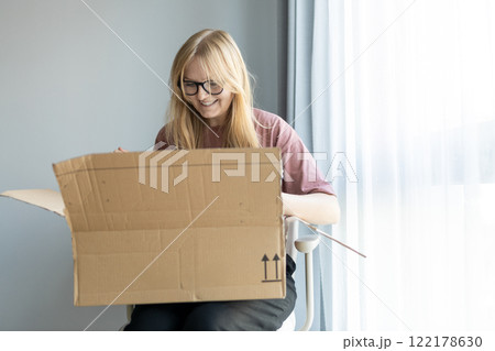 Young cheerful woman in glasses getting parcel at home. Pretty girl sitting on sofa at home and opening carton box. Express delivery service and satisfied consumer. Online shopping banner 122178630