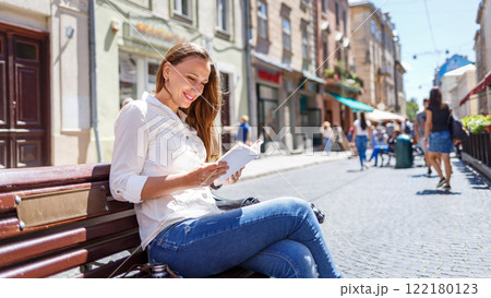 A young woman reading on a sunny day sitting on a bench in a lively street filled with pedestrians and vibrant shops 122180123