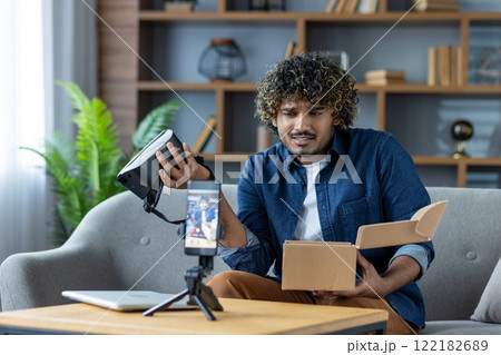 Man writes negative review about product purchased in online store. Influencer blogger sits at home on sofa in living room, uses tripod and phone to record online video stream. Man writes negative review about product purchased in online store. Influencer blogger sits at home on sofa in living room, uses tripod and phone to record online video stream. 122182689