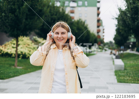 Smiling young 30s woman listening to music through wireless headphones on urban background. Music lover enjoying music, chill moment Smiling young 30s woman listening to music through wireless headphones on urban background. Music lover enjoying music, chill moment 122183695