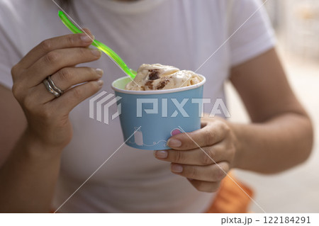 a woman in a white T-shirt eats ice cream from a paper cup with a spoon, summer, heat a woman in a white T-shirt eats ice cream from a paper cup with a spoon, summer, heat 122184291