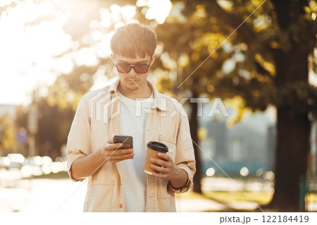 Smiling Young Man Texting on Smartphone and Holding Coffee in Morning City Street 122184419