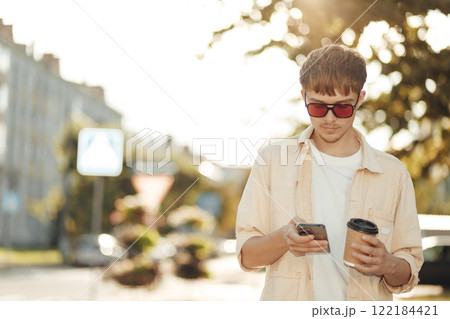 Smiling Young Man Texting on Smartphone and Holding Coffee in Morning City Street Smiling Young Man Texting on Smartphone and Holding Coffee in Morning City Street 122184421