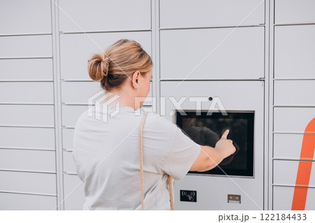 Parcel delivery machine. Mail delivery and post service concept. Happy smiling woman with phone at outdoor automated parcel machine. Electronic locker for storing parcels. Parcel delivery machine. Mail delivery and post service concept. Happy smiling woman with phone at outdoor automated parcel machine. Electronic locker for storing parcels. 122184433