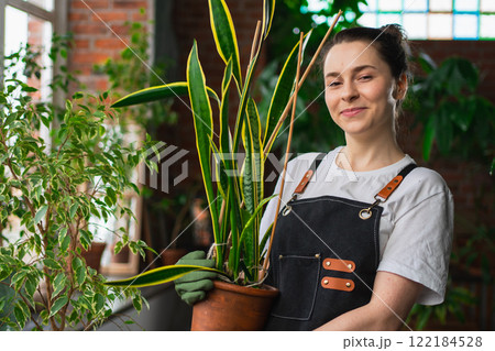 Running of own business. Young woman florist holding plant in pot wearing apron in botanical store. Happy small business owner working at flower shop smiling surrounded by plants. Small business 122184528
