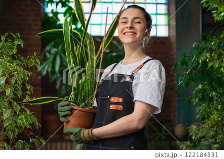 Running of own business. Young woman florist holding plant in pot wearing apron in botanical store. Happy small business owner working at flower shop smiling surrounded by plants. Small business 122184531