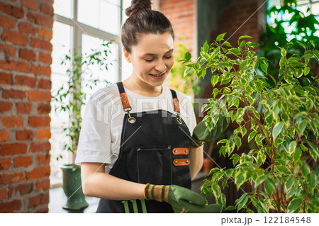 Running of own business. Woman florist wearing apron taking care of green plants in botanical store. Happy small business owner working at flower shop standing surrounded by plants. Small business 122184548