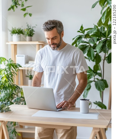Young man in casual clothing working on a laptop at a plant-filled desk, bright and airy environment, concepts of modern work and minimalistic workspace design 122185200