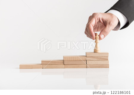Businessman placing queen chess piece on a stepped stack of wooden blocks Businessman placing queen chess piece on a stepped stack of wooden blocks 122186101