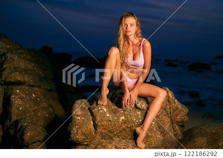 Stunning model in bikini sits on ocean beach rocks at night. Sensual woman poses in evening light. Perfect for dating apps or fashion ads. Captures allure, attraction, and feminine charm. Stunning model in bikini sits on ocean beach rocks at night. Sensual woman poses in evening light. Perfect for dating apps or fashion ads. Captures allure, attraction, and feminine charm. 122186292