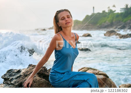 Blonde model in wet dress on cliff with ocean waves and exotic island. Her elegance embodies divine beauty and femininity in nature. Perfect for fashion, travel, nature, or lifestyle themes. 122186302