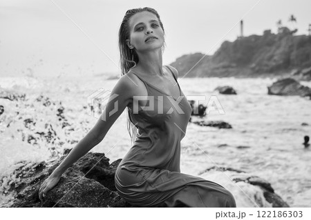 Young woman in wet dress by ocean rocks, feels free. Her look is serene, captures divine beauty. Waves crash, nature vibes, femininity, elegance. Black and white style, timeless allure, summer day. 122186303