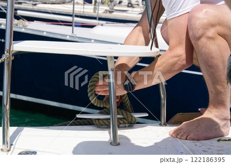Close-up of a man s hand untying a rope on a cleat on a boat deck at the marina while barefoot. Close-up of a man s hand untying a rope on a cleat on a boat deck at the marina while barefoot. 122186395