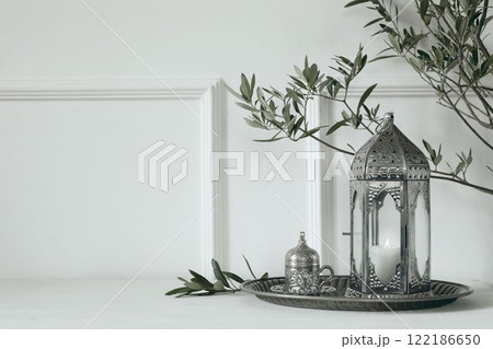 Ramadan Kareem holiday still life. Ornamental silver Moroccan lantern with olive tree branches on wooden table. Cup of Turkish tea. Blurred white wall background. Eid Mubarak, Eid al Adha holiday Ramadan Kareem holiday still life. Ornamental silver Moroccan lantern with olive tree branches on wooden table. Cup of Turkish tea. Blurred white wall background. Eid Mubarak, Eid al Adha holiday 122186650