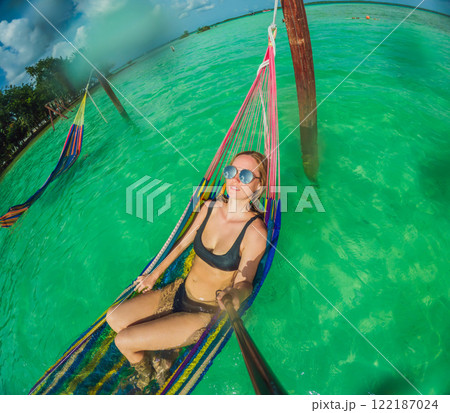 Woman tourist relaxing on a hammock above the turquoise waters of Lake Bacalar, Mexico. Peaceful retreat, tropical paradise, and relaxation concept in a serene natural setting Woman tourist relaxing on a hammock above the turquoise waters of Lake Bacalar, Mexico. Peaceful retreat, tropical paradise, and relaxation concept in a serene natural setting 122187024