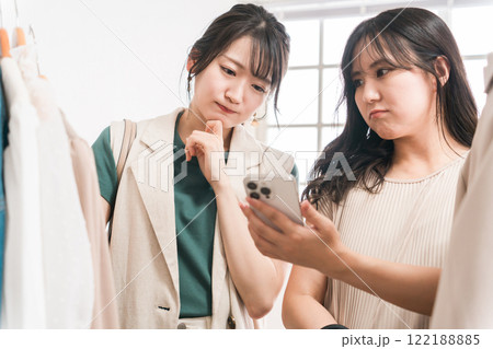 洋服屋・アパレルショップでスマホを見ながら洋服を選ぶ女性・友達（買い物・ショッピング） 122188885