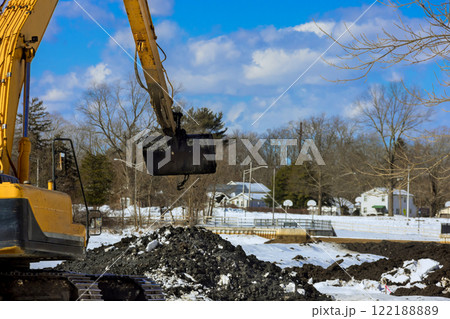 An excavator operates in snowy environment, moving earth near homes under works foundation area 122188889