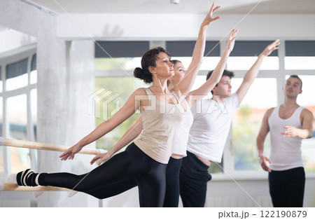 Pretty female ballerina practicing arabesque pose. Young male ballet trainer teaching group of dancers by arabesque pose near ballet barre in dancing hall 122190879