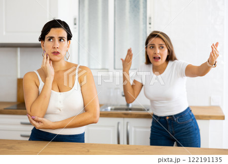 Frustrated young woman listening to angry girlfriend in home kitchen 122191135