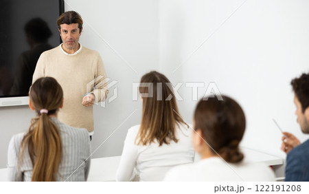 Male trainer near interactive board conducting advanced training courses to office employees sitting at desks in auditory 122191208